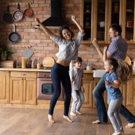 Barefoot dances. Overjoyed millennial family with little daughter son having fun listening to music dancing. Active mom dad and two junior school age kids jump at kitchen celebrate moving to new home